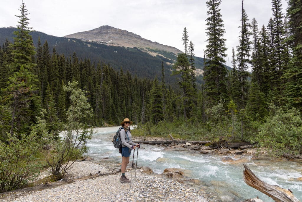 Woman holding trekking poles standing next to river on hike in British Columbia