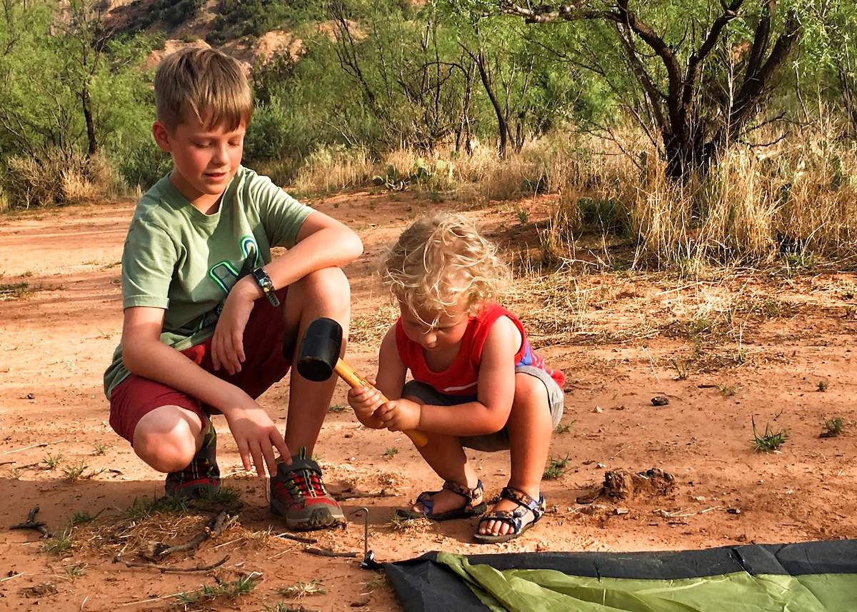 Child hammering tent stake into ground with rubber hammer and older child supervising