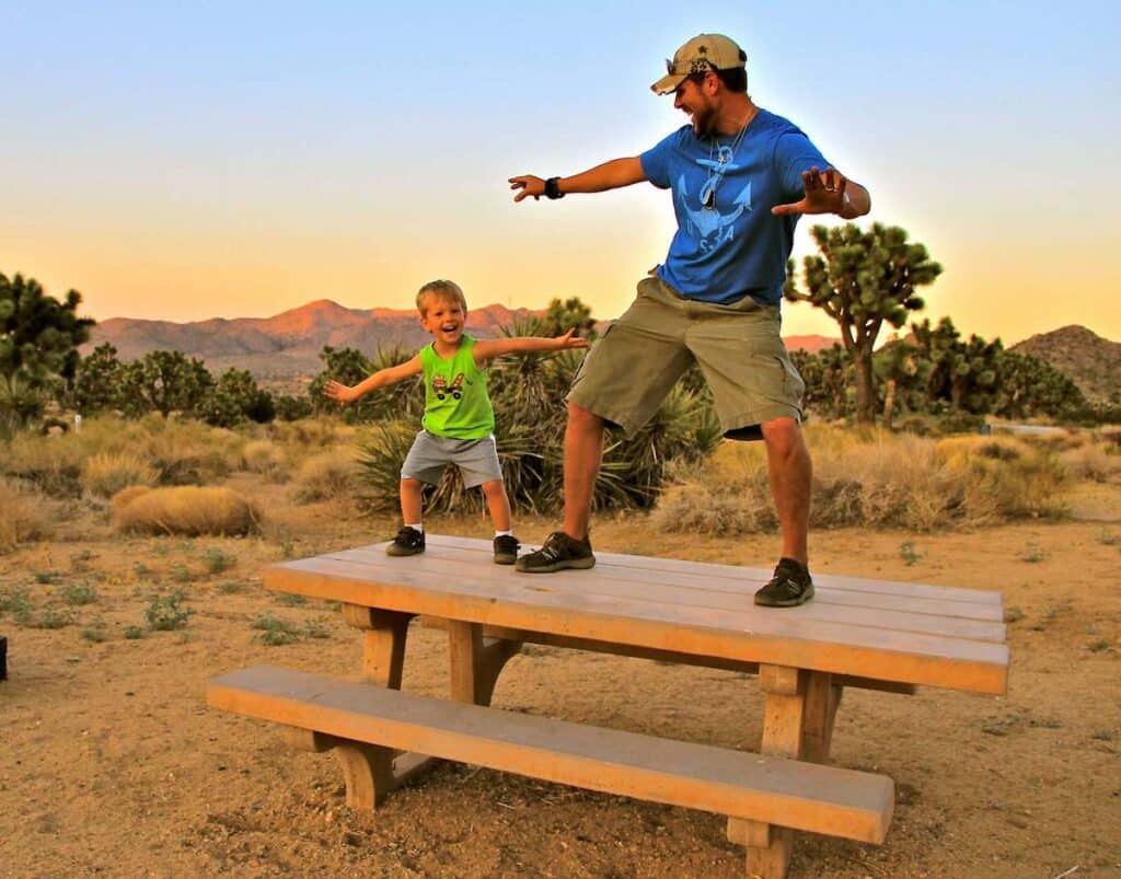 Man and son standing on picnic table at desert campground pretending to surf with sunset in background 