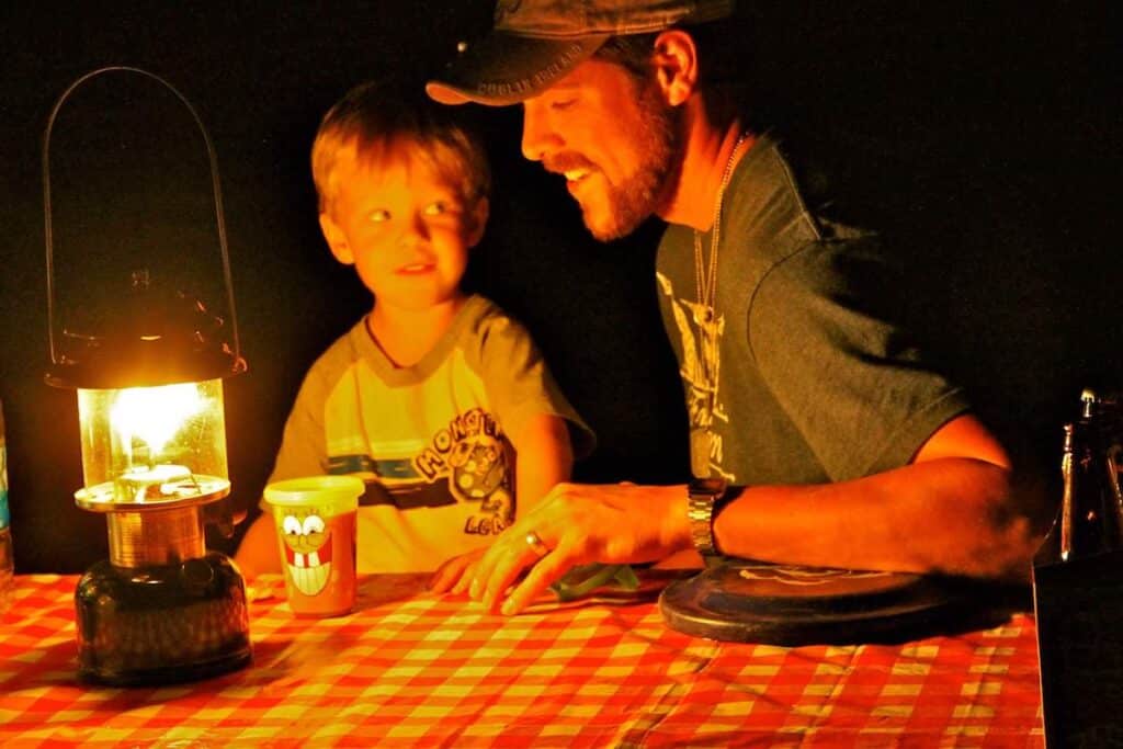 man sitting with child at picnic table at campground at night with light by Coleman Gas Lamp