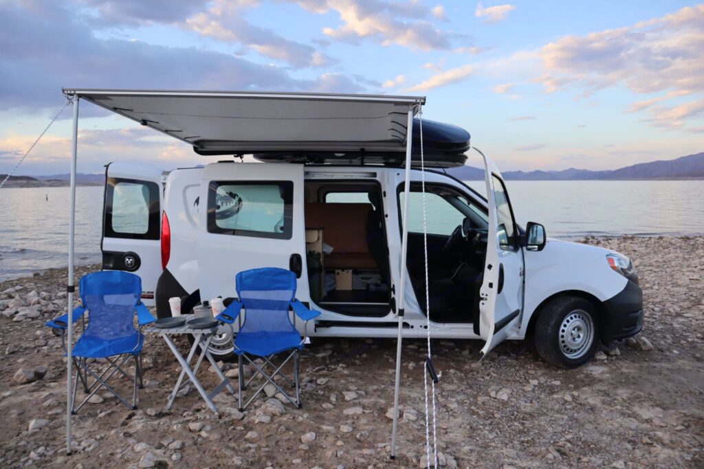 The side of a micro-camper from VegasCampers.com with an awning extended and 2 camp chairs set up outside