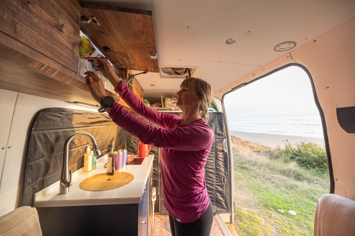 Women in a camper van reaching in her cabinets with the door open and a view of the ocean