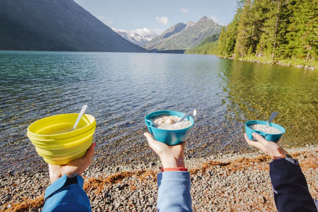 Three arms holding bowls of porridge next to lake on backpacking trip