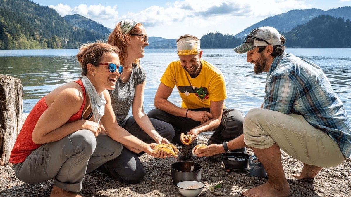 Four people squatting around camp stove by lake holding loaded tacos on a backpacking trip