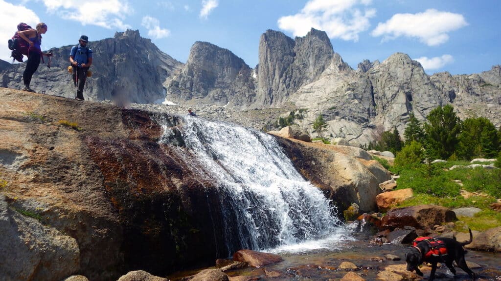 two hikers stand next to a waterfall on Cirque of the Towers backpacking trip.