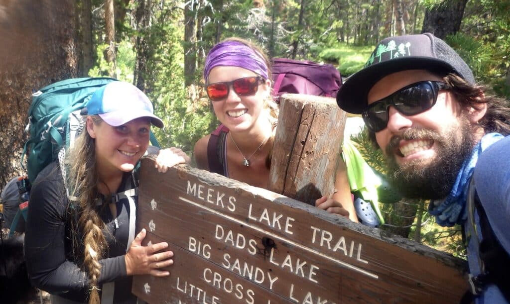 three backpackers smile at the Cirque of the Towers sign on Meeks Lake Trail