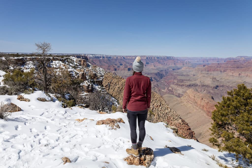 Woman wearing a fleece, leggings, beanie, and hiking boots looking out over the Grand Canyon in the snow