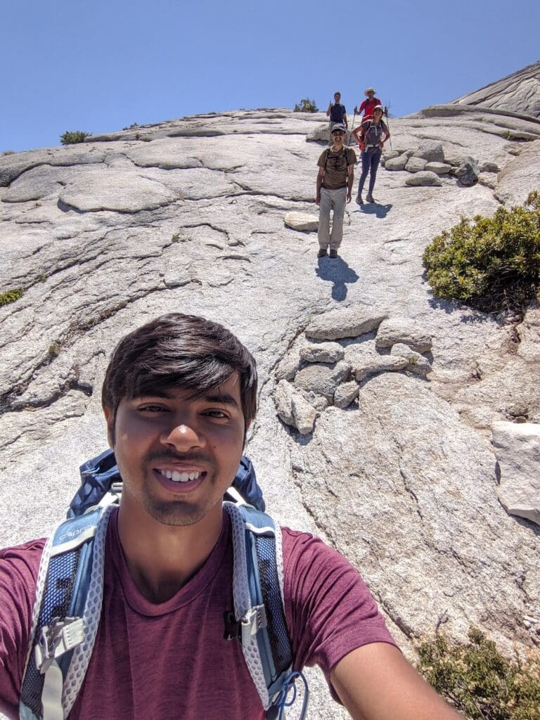 A man takes a selfie on the subdome of Half Dome with friends smiling in the background at a distance