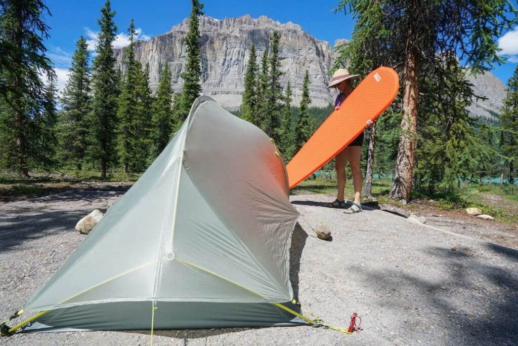 Man inflating sleeping pad at campsite with tent set up and tall rocky mountain in background