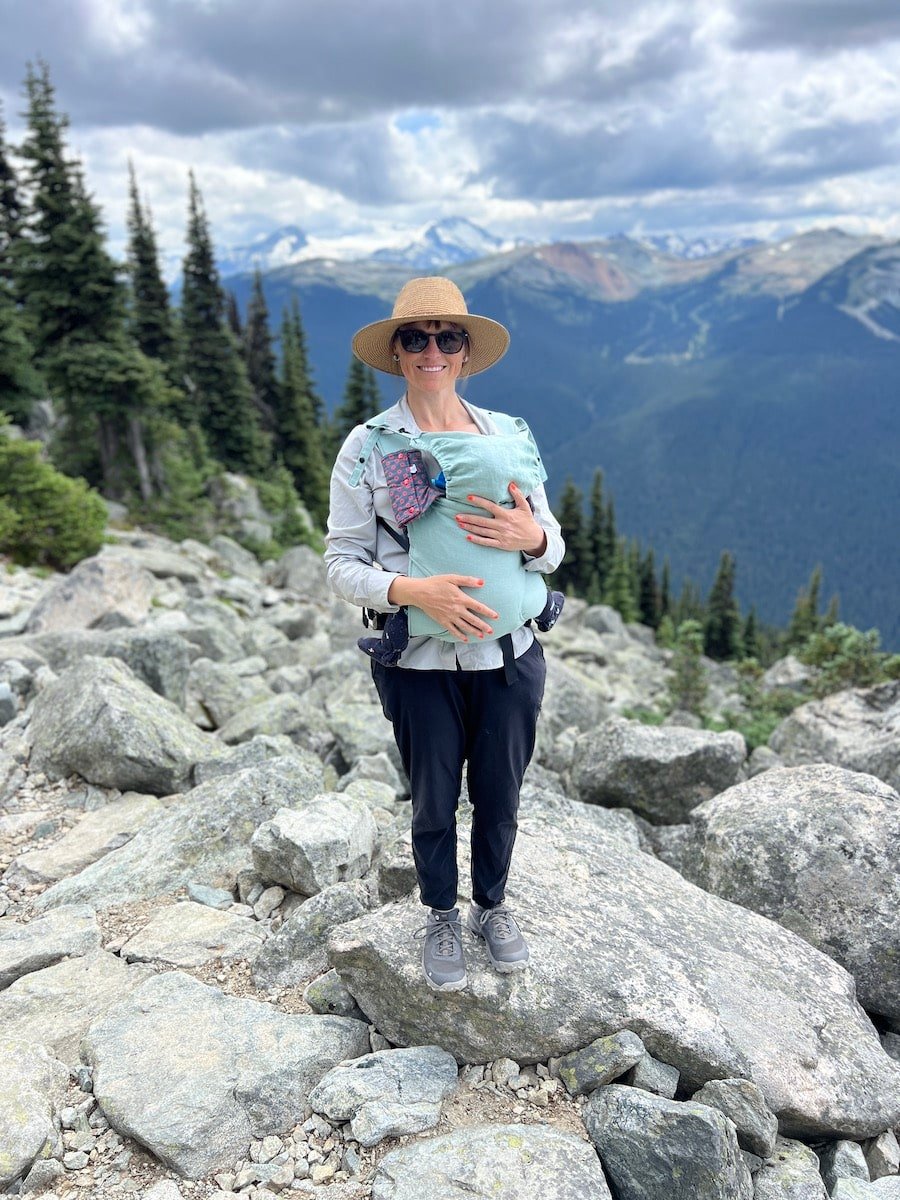 Kristen Bor standing on a rock holding a baby in a baby carrier with the view of the mountains around Whistler in the background