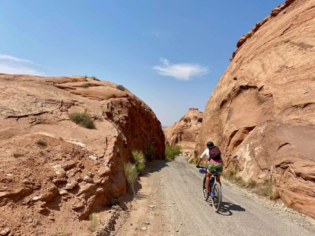 Mountain biker riding bike down Sand Flats Road in Moab. Tall red rock boulders on either side of the road