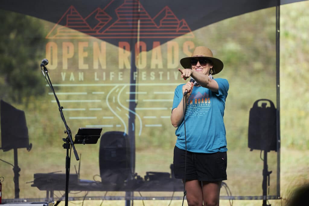 Bearfoot Theory and Open Roads Fest founder Kristen Bor speaking at opening night of Open Roads Fest. There is a banner with the Open Roads Fest logo behind her