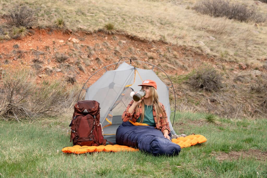 Photo of a woman sitting outside her tent on a sleeping pad with the REI Magma sleeping bag