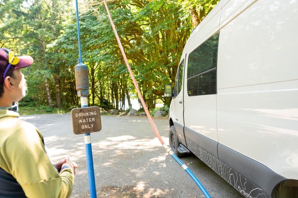 Man standing next to potable water spigot filing up the tanks in a Sprinter Van with a hose