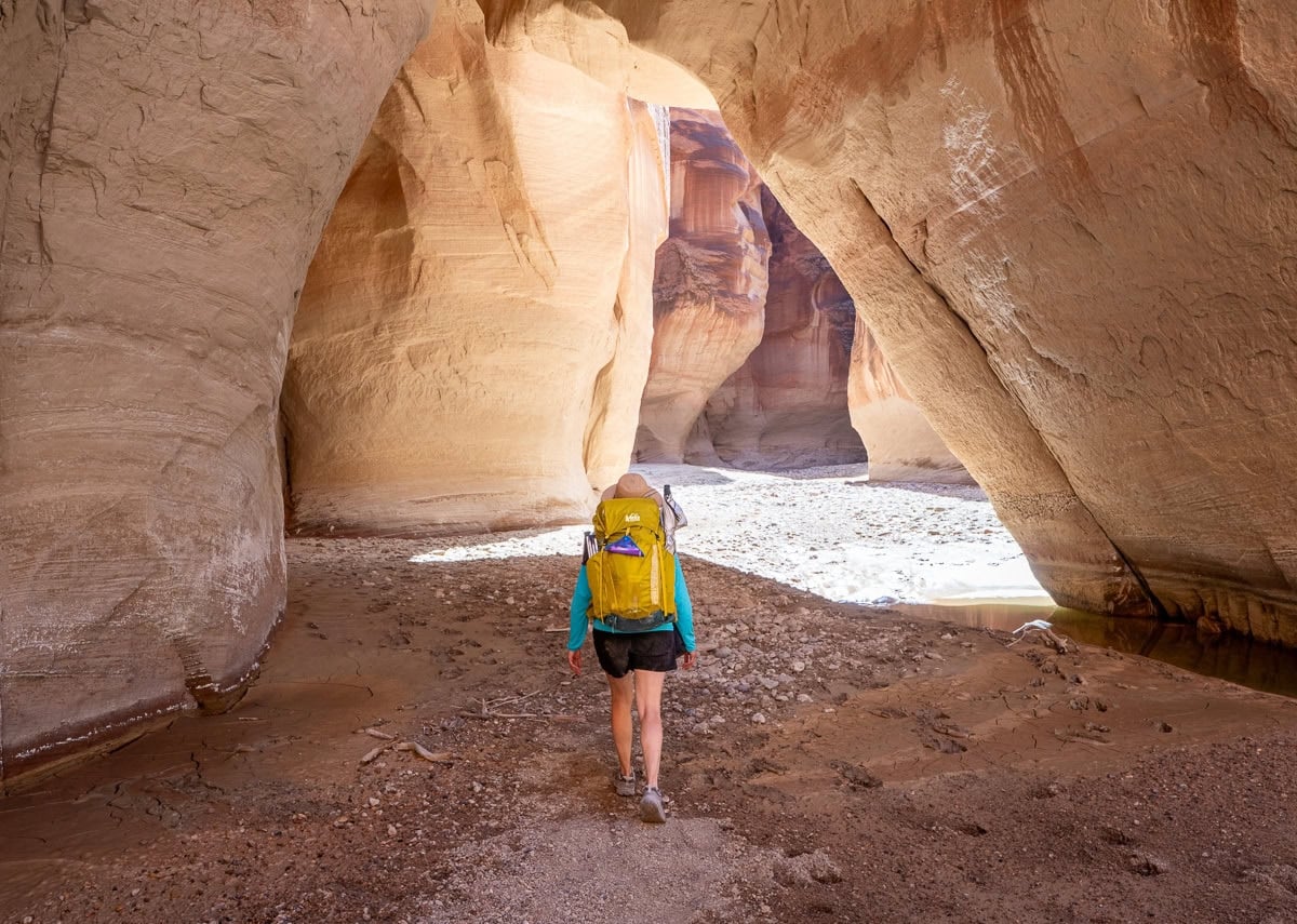 The back of a woman wearing a yellow REI backpacking pack walking through a red rock tunnel in a river bed